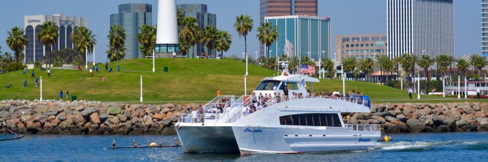 White passenger catamaran leaving a harbor, with a green park and city skyline behind